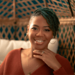 Smiling person with green locs and orange sweater.