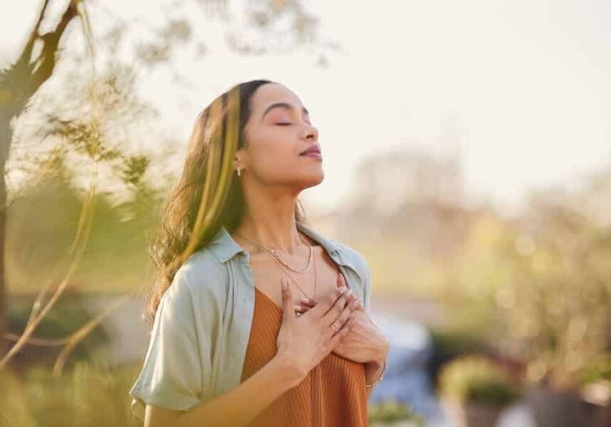 Young latin woman with hand on chest breathing in fresh air in a beautiful garden during sunset. Healthy mexican girl enjoying nature while meditating during morning exercise routine with closed eyes. Mindfulness woman enjoying morning ritual and relaxing technique.